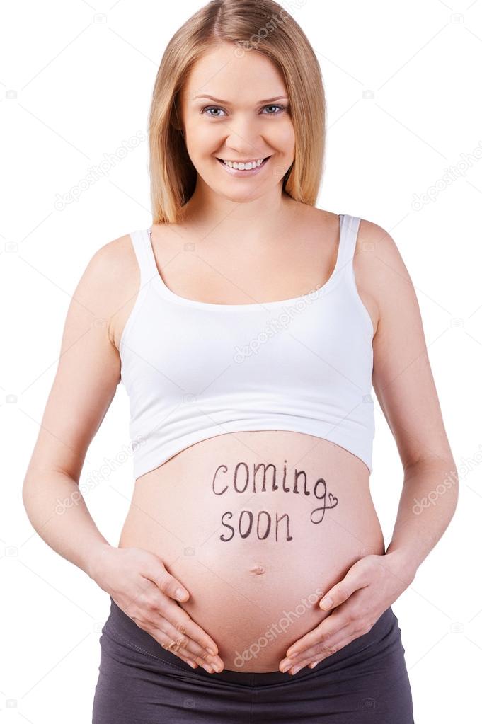 Coming soon. Cropped image of pregnant woman with coming soon sign on her belly standing isolated on white