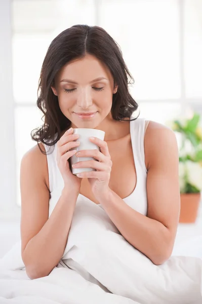 Détente avec une tasse de café frais. Attrayant jeune femme souriante assis dans le lit et tenant une tasse de café — Photo