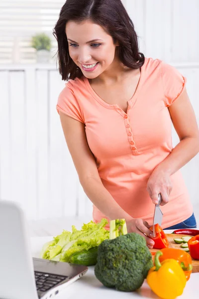 Kvinna som tittar på laptop och skära grönsaker — Stockfoto