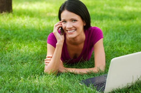 Dizüstü bilgisayar ile çim yatan telefonda konuşan kadın — Stok fotoğraf