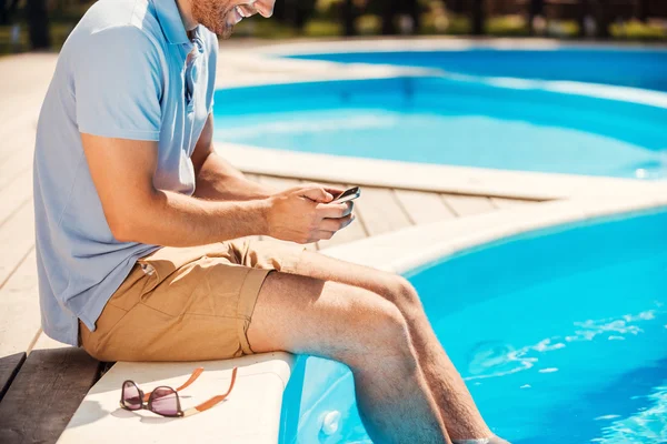 Homem sentado à beira da piscina e digitando no celular — Fotografia de Stock