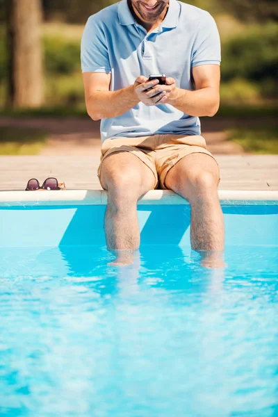 Homem sentado à beira da piscina e digitando no celular — Fotografia de Stock