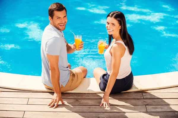 Casal segurando copos com suco e sentado à beira da piscina — Fotografia de Stock