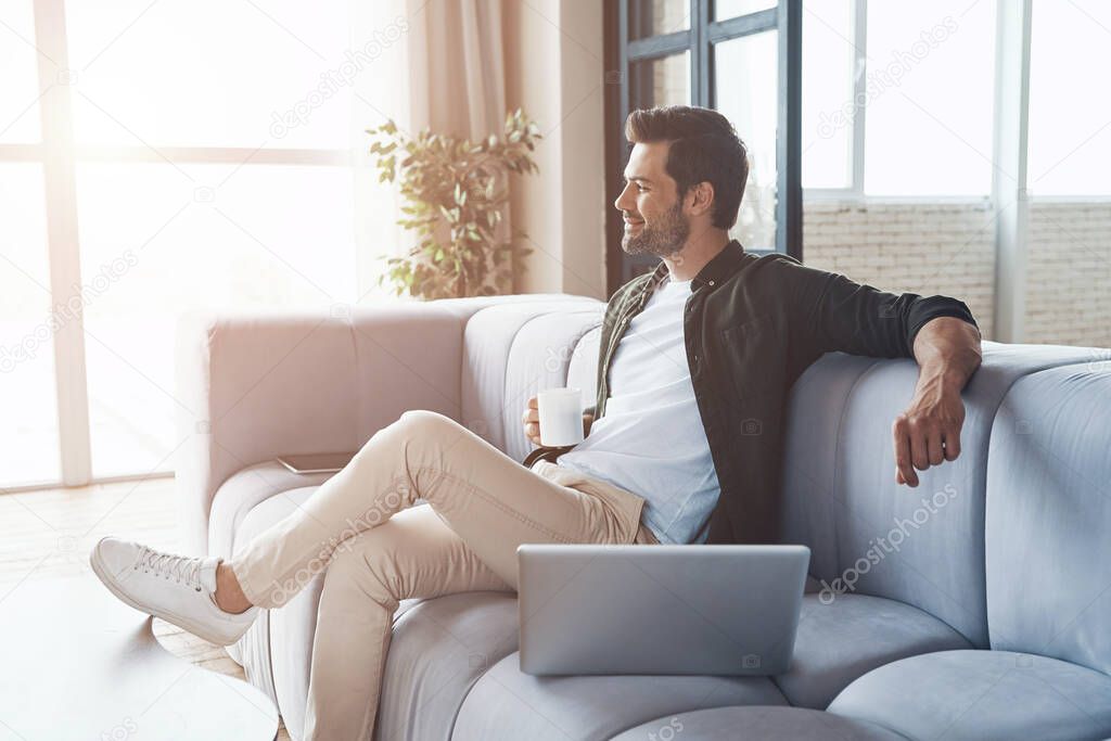 Thoughtful young man drinking coffee and looking away while sitting on the sofa