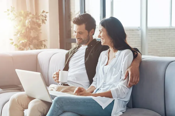 Mooi jong stel drinken van koffie en kijken op de laptop terwijl zitten op de bank thuis — Stockfoto