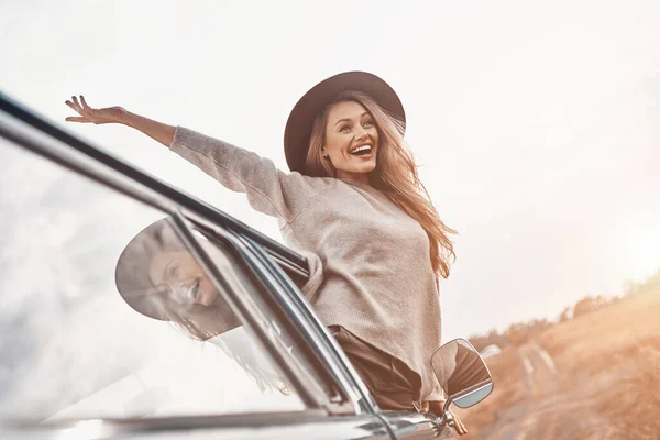 Vacker ung kvinna i hatt hålla armarna utsträckta och ser glad ut medan du njuter av road trip i minivan — Stockfoto
