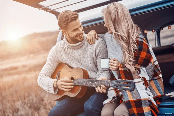 Guapo joven tocando la guitarra para su novia mientras ambos están sentados en el maletero del coche al aire libre —  Fotos de Stock