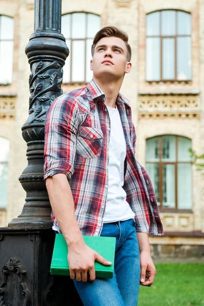 Student holding book and standing against university building — Stock Photo, Image
