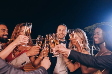 Formalite icabı giyinmiş bir grup insan şampanyayla kadeh kaldırıyor ve lüks partilerde vakit geçirirken gülümsüyor.