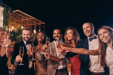 Formalite icabı giyinmiş bir grup insan şampanyayla kadeh kaldırıyor ve lüks partilerde vakit geçirirken gülümsüyor.