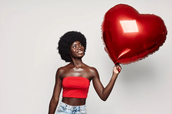 Beautiful young African woman holding red heart shape balloon and smiling — Stock Fotó