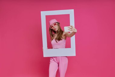 Pembe arka planda dikilirken resim çerçevesine bakan oyuncu genç kadın.