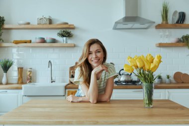 Mutfak masasına yaslanıp gülümseyen mutlu genç kadın.