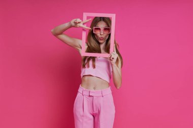 Pembe arka planda dikilirken resim çerçevesine bakan oyuncu genç kadın.