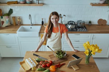 Genç ve güzel bir kadının kameraya bakıp mutfakta yemek pişirirken gülümsemesi.