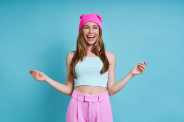 Mavi arka planda dururken gülümseyen pembe şapkalı mutlu genç kadın.