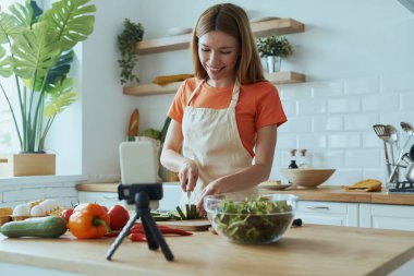 Güzel, genç bir kadın ev mutfağında yemek pişiriyor ve video kaydediyor.