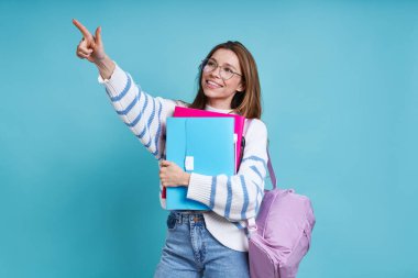 Mutlu kız öğrenci mavi arka planda dikilirken işaret ediyor