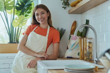 Güzel genç bayan mutfak lavabosunun yanında duruyor ve gülümsüyor.