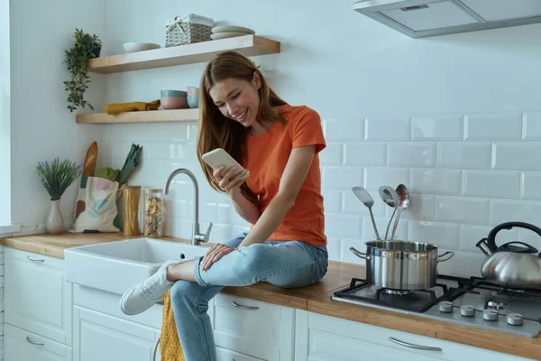 Attractive young woman using smart phone while sitting at the kitchen counter