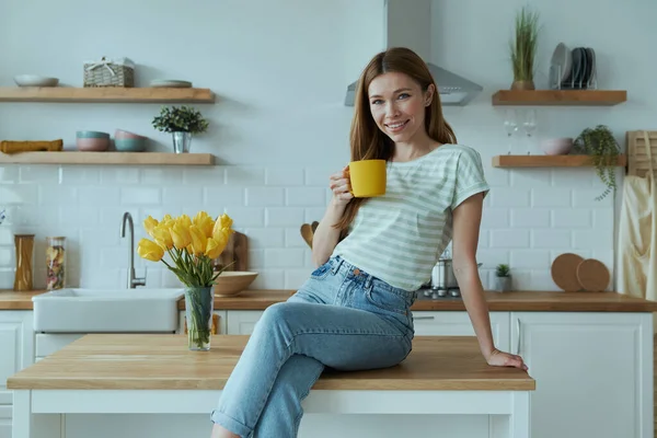 Cheerful young woman holding yellow cup and looking at camera while sitting on the kitchen counter