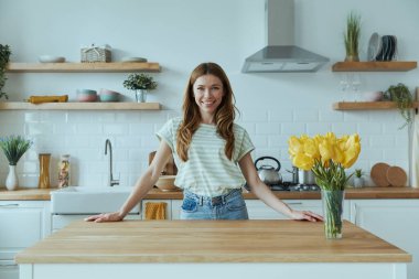 Güzel genç bir kadın mutfak masasına yaslanıyor ve gülümsüyor.