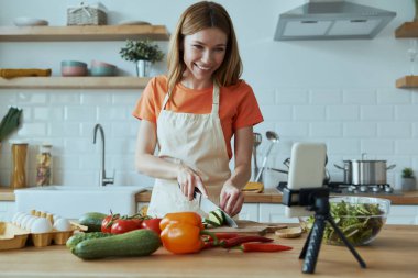 Çekici genç bir kadın ev mutfağında dururken yemek pişiriyor ve video kaydediyor.