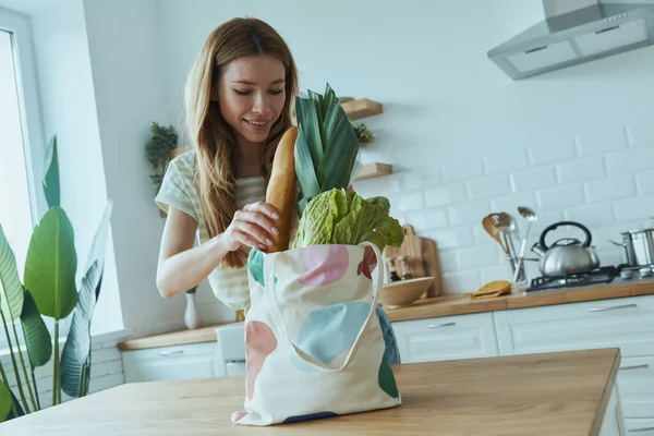 Happy young woman unpacking the bag with healthy food while standing at the domestic kitchen