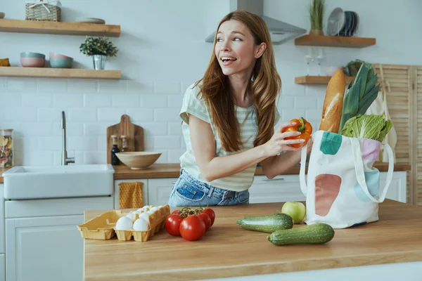 Beautiful young woman unpacking the bag with healthy food while standing at the domestic kitchen