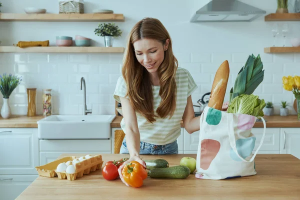 Happy young woman unpacking the bag with healthy food while standing at the domestic kitchen
