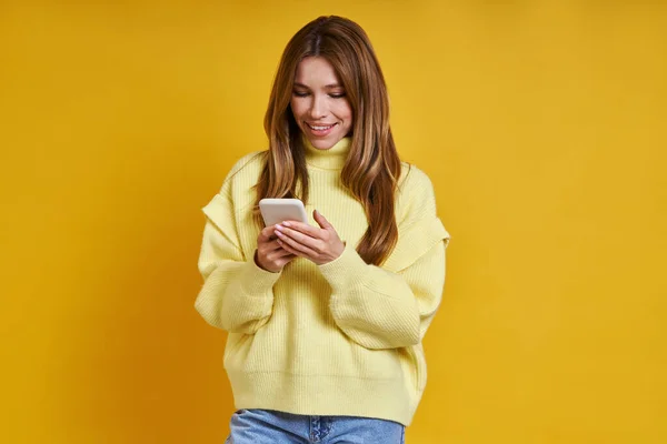 Счастливая Молодая Женщина Используя Свой Смартфон Стоя Желтом Фоне — стоковое фото