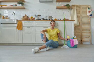 Mutfaktaki temizlik malzemeleriyle kovanın yanında oturan neşeli genç kadın.