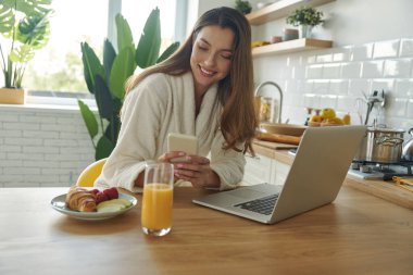 Modern mutfakta vakit geçirirken bornozlu güzel bir kadın teknoloji kullanıyor.