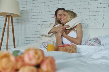 Güzel bir çift yatakta kucaklaşırken bir demet gül ön planda yatıyor.