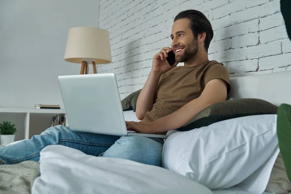 Jovem Alegre Usando Laptop Falando Celular Enquanto Relaxa Cama Casa — Fotografia de Stock