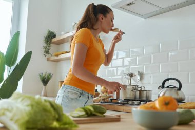 Mutfakta yemek pişirirken tavadan bir şeyler tadıyor.