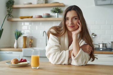 Modern mutfakta vakit geçirirken kameraya bakan bornozlu çekici genç bir kadın.