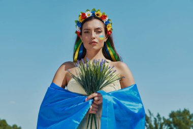 Güzel Ukraynalı kadın elinde çiçeklerle arka planda gökyüzü olan kameraya bakıyor.