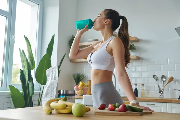 Beautiful young woman in sports clothing drinking protein cocktail while standing at the kitchen