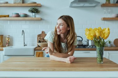 Güzel genç bir kadın mutfak masasına yaslanıyor ve gülümsüyor.