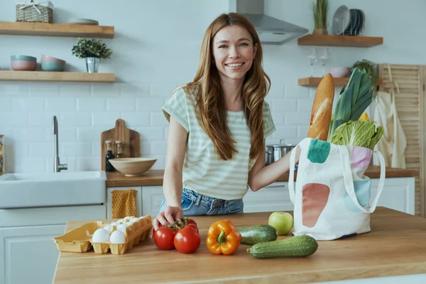 Beautiful young woman unpacking the bag with healthy food while standing at the domestic kitchen