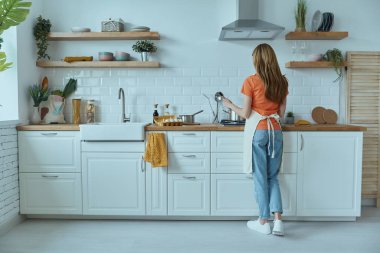 Genç bir kadının ev mutfağında yemek pişirişinin tam boy dikiz görüntüsü.