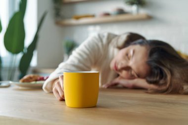 Bornozlu genç bir kadın evdeki mutfak tezgahında otururken bir fincan kahveyle kestiriyor.