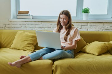 Dizüstü bilgisayarda çalışma ve evde kanepede otururken gülümseyen güzel genç kadın