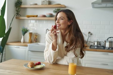 Bornozlu genç kadın modern mutfakta kahvaltı yapıyor.