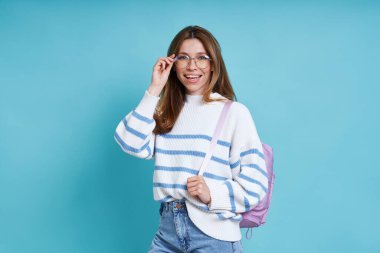 Mavi arka planda dururken sırt çantasıyla gözlük takan mutlu kız öğrenci