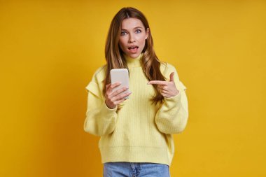 Şaşırmış genç bir kadın sarı arka planda dururken akıllı telefonunu işaret ediyor.