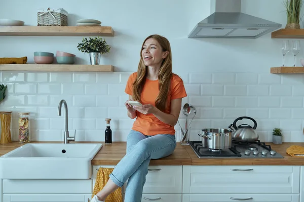 Güzel genç bayan mutfak tezgahında otururken akıllı telefon kullanıyor.