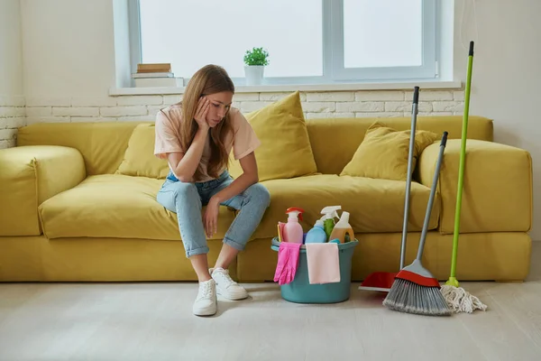 Bored woman looking at the bucket with cleaning products while sitting on the couch at home