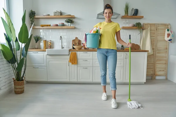 Güzel genç bir kadın mutfakta dururken paspas ve temizlik malzemesi taşıyor.